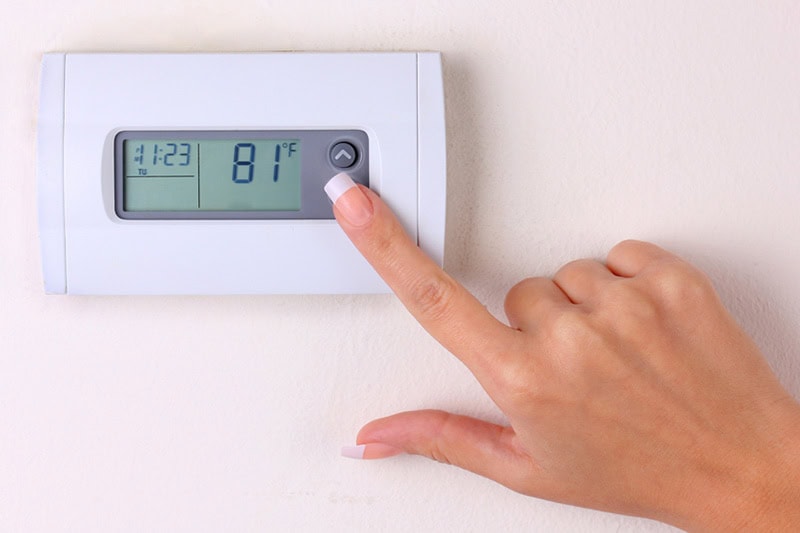 Thermostat 101: Basic Buyer’s Guide. Photo of a person adjusting a digital thermostat mounted on a wall.